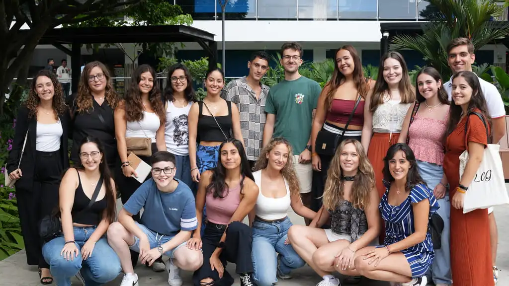 Fotografía grupal de 18 personas, todos integrantes de la Universidad de Córdoba, España. Es un grupo compuesto por jóvenes adultos, posando al aire libre. Están ubicados en “El Bosque”, un ambiente verde y agradable, del campus universitario de la USC, con vegetación en el fondo y un edificio de varios pisos visible detrás de ellos. Los rostros muestran sonrisas, reflejando un momento de camaradería y alegría.