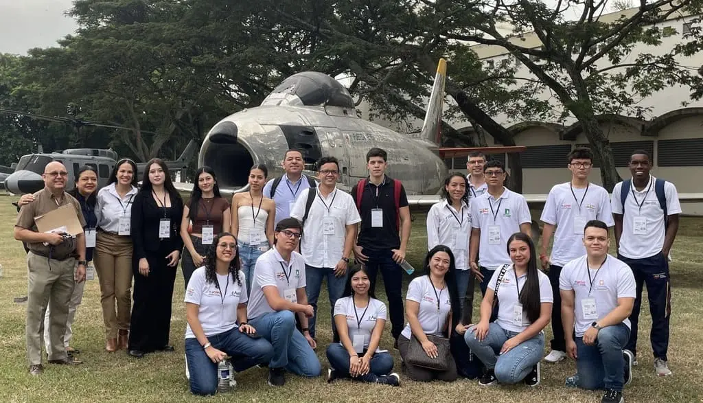Grupo de personas, en su mayoría jóvenes con camisetas blancas y gafetes, posando frente a un avión militar antiguo al aire libre, rodeados de árboles.
