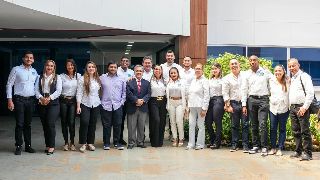 Grupo de personas, posando para una foto grupal frente a un edificio moderno con ventanales grandes. Todos llevan camisas formales blancas o de colores claros. La escena está iluminada por luz natural.