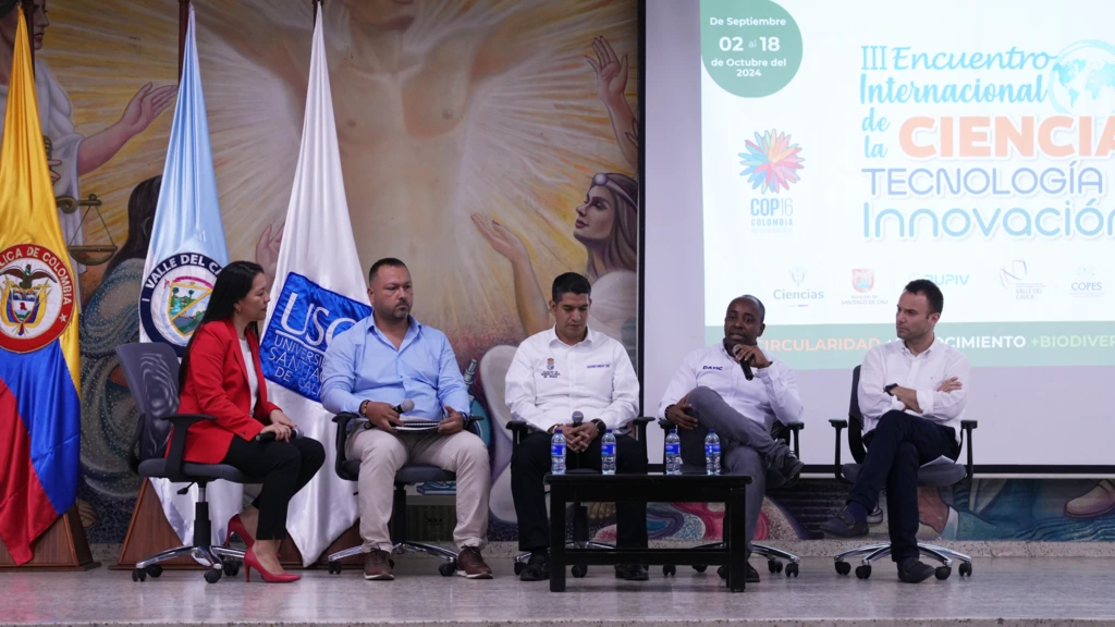 Un grupo de cinco personas está sentado en un escenario en frente de un público, durante un panel de discusión en un evento llamado "III Encuentro Internacional de la Ciencia, Tecnología e Innovación". Las personas son cuatro hombres y una mujer. Detrás de ellos hay banderas de Colombia, del Cauca, y logos de varias universidades y organizaciones. Un mural colorido de temática científica y tecnológica adorna el fondo.