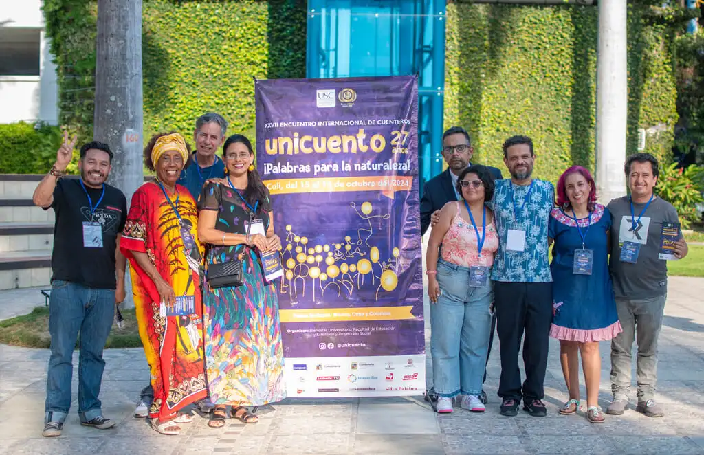Participantes posan sonrientes frente a un cartel que anuncia el XXVII Encuentro Internacional de Cuenteros Unicuento 2024 con el lema "¡Palabras para la naturaleza!". El entorno muestra vegetación, transmitiendo una atmósfera alegre y cultural.
