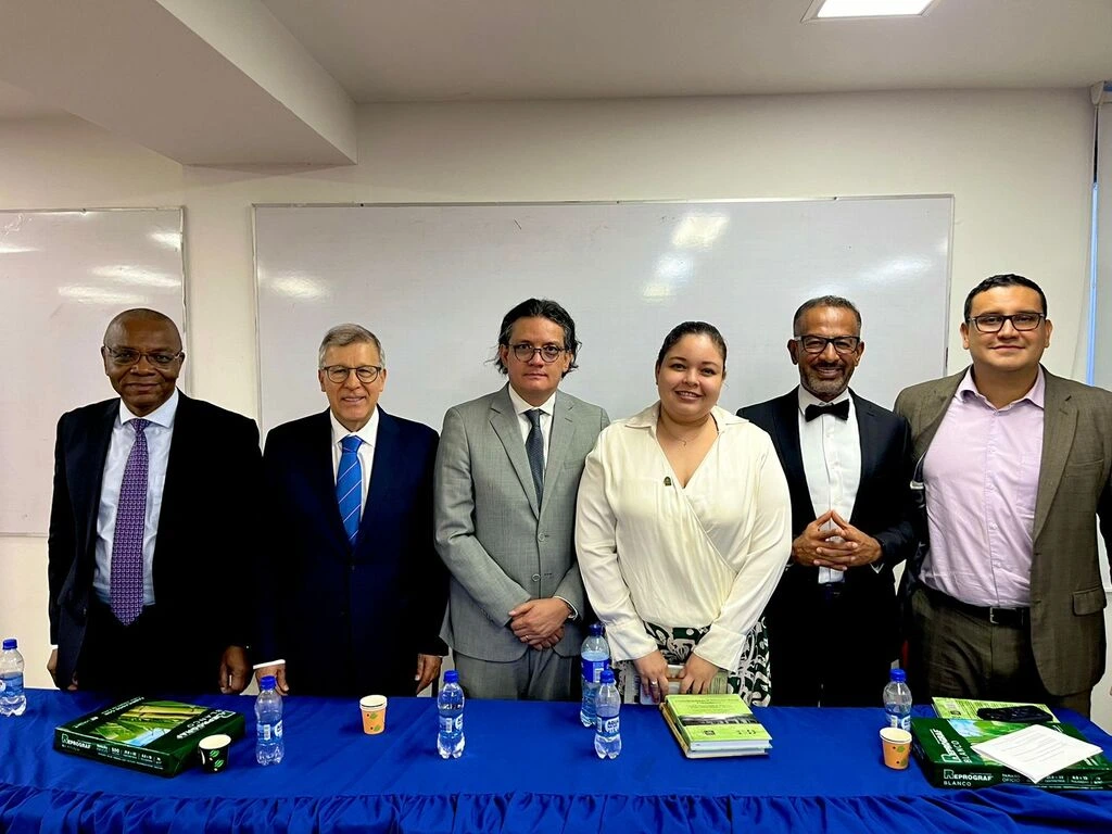 Seis personas están de pie detrás de una mesa azul en una sala de reuniones. Los hombres y mujeres, vestidos de manera formal, están posando para una foto grupal frente a una pizarra blanca. En la mesa hay botellas de agua, documentos y materiales de conferencia.