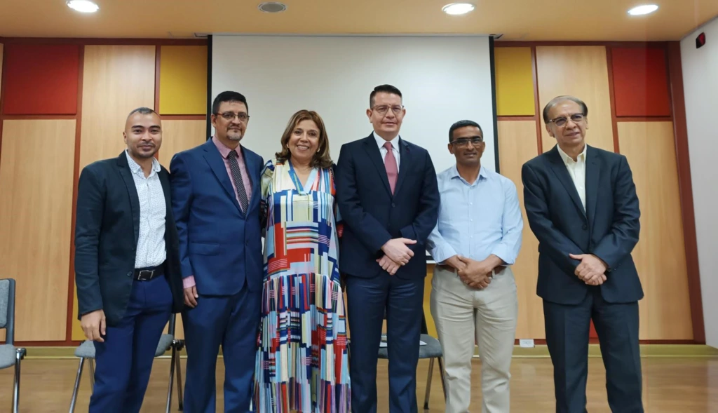 Seis personas posan de pie y sonríen hacia la cámara en un auditorio con paneles de colores cálidos en el fondo. Están vestidos formalmente y parecen participar en un evento académico o institucional.